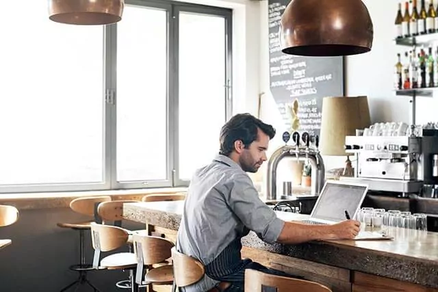 Man working on a laptop and jotting notes at a bar counter in a cozy cafe, with large windows in the background.