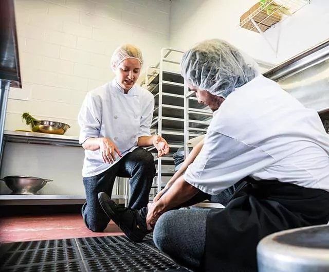 Kitchen worker knelt on the floor and holding their leg in pain, with another colleague offering assistance.