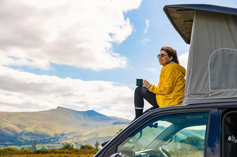 Person with a mug on a vehicle roof tent, enjoying a scenic landscape, with a mountain and a cloudy sky in the background.