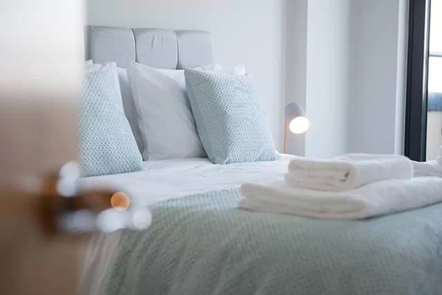 Neatly made bed in a cozy room with grey headboard, blue pillows, and white towels on the bed.