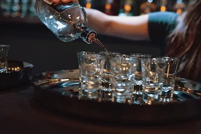 Bar staff pouring shots into glasses on a metal tray, in a softly lit bar.
