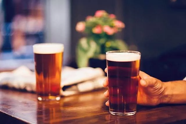Hand holding a pint of beer on a table, with another glass, newspaper, and flowers on the table.