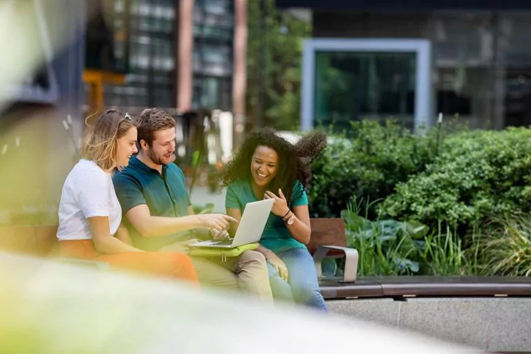 Group of three professionals having a discussion outdoors at a table, with greenery and buildings in the background.