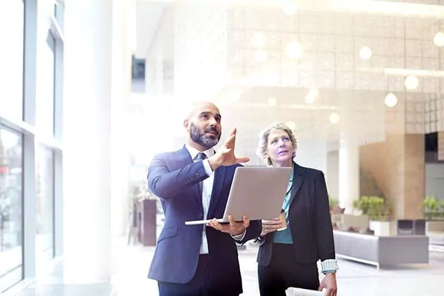 A man holding an open laptop and a woman, both in business attire, standing in a very bright room.