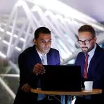 Two businessmen are looking at a laptop in a dimly lit, modern office setting.