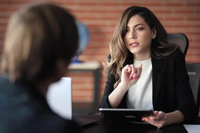 A woman in a business suit engages in conversation with someone else while holding a tablet in an office setting.