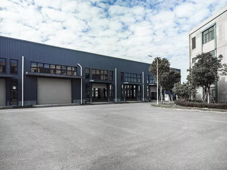 Industrial building with blue panelling, garage doors, and greenery, under a cloudy sky.