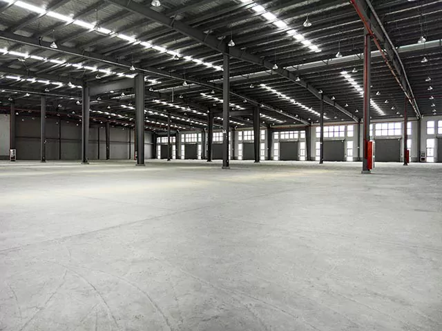 Spacious, empty warehouse with concrete floor, metal beams, and natural light coming in from big windows.