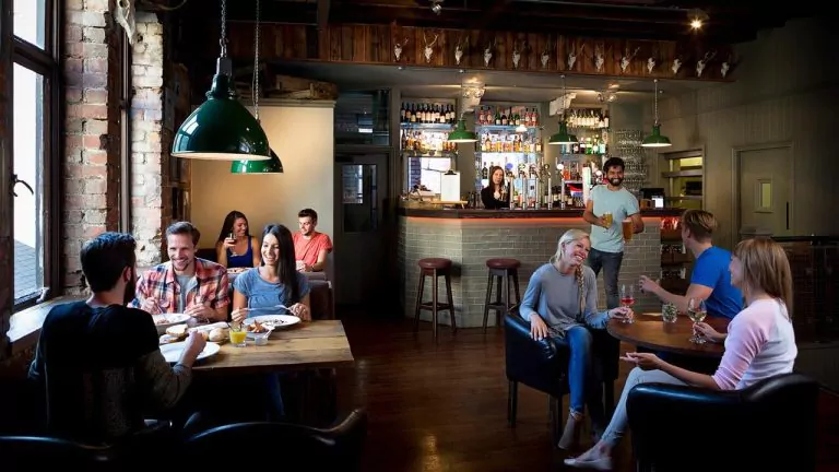Cozy restaurant with wooden tables, groups dining, and a bartender serving drinks in a warm, rustic setting.