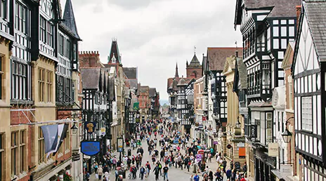A bustling street scene in a historic town, featuring Tudor-style buildings with black and white timber framing. The street is filled with people walking, and the sky is overcast, adding a subdued atmosphere to the scene.