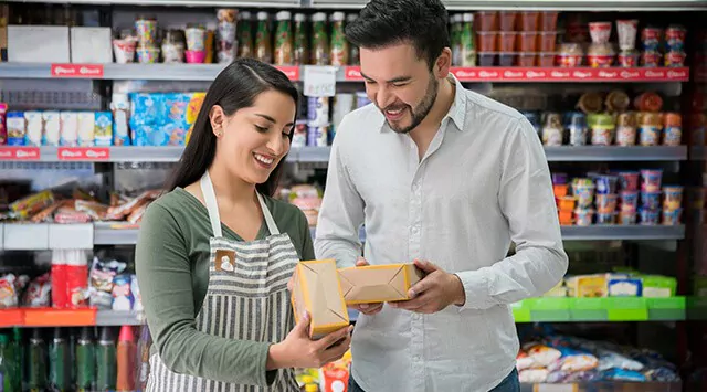 A convenience store worker in an apron and a man, smiling while holding packaged food items.