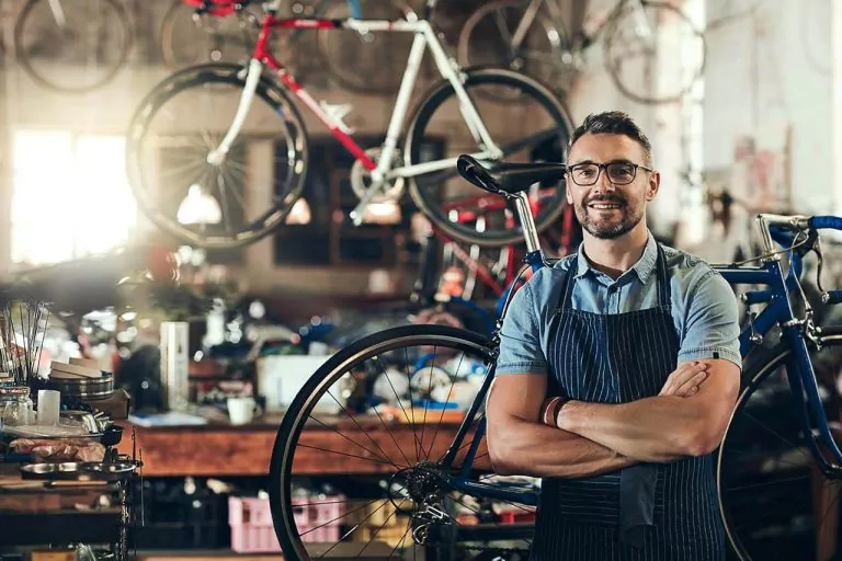 Man in an apron with arms crossed in a bicycle workshop, surrounded by bikes and tools.
