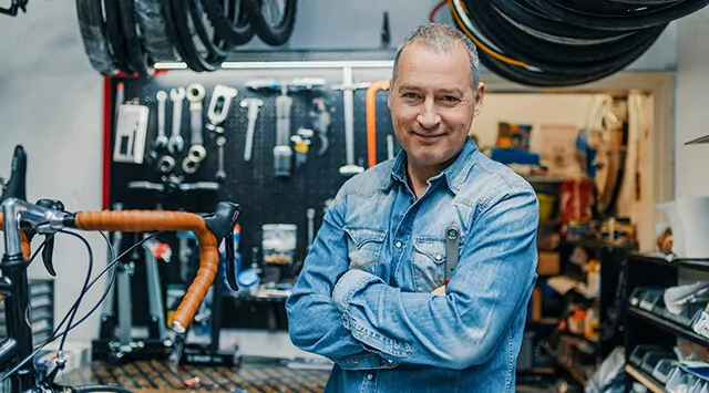 A man in a denim shirt stands confidently in a bicycle repair shop. He is surrounded by bike tools hanging on the wall and various bike parts. A bicycle with brown handlebar tape is partially visible in the foreground.