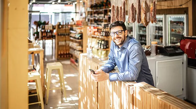 Man leaning on a counter in a delicatessen shop, smiling and holding a smartphone.