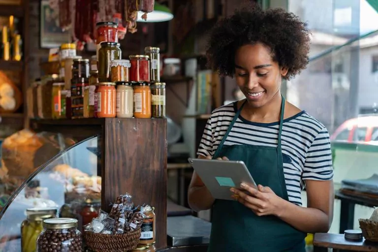 Smiling deli worker using a tablet, surrounded by shelves of jars.