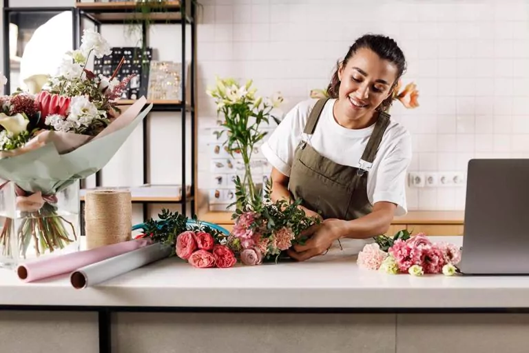 Florist arranging a bouquet on a counter, with gift wrapping materials and decorations in the background.