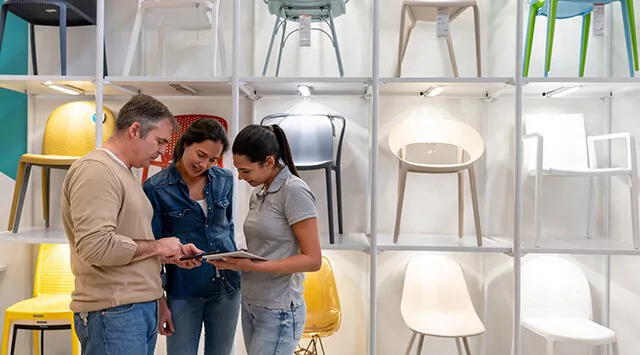 Three people in a furniture shop are standing by a shelving unit with chairs, looking at a tablet that one of them is holding.