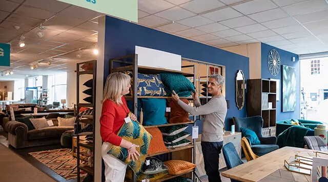 Man and woman selecting cushions in a furniture store, surrounded by couches and decor items.
