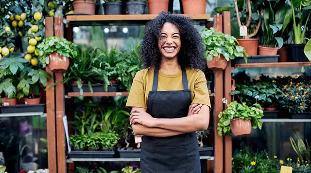 Person wearing and apron, smiling with arms crossed in a garden center, surrounded by potted plants and flowers.