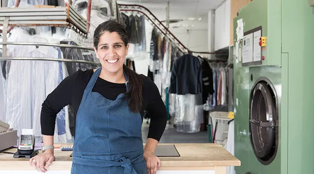 A launderetter owner wearing a blue apron smiles in front of the counter with clothing and machines in the background.
