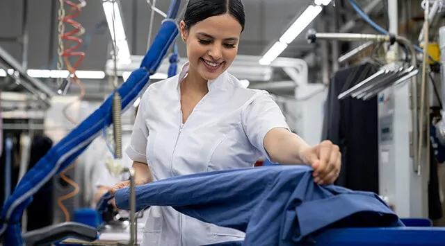Woman wearing a white uniform working in a laundry facility, holding a shirt and operating a pressing machine.