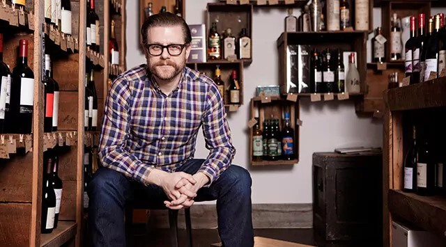 A man wearing glasses and a plaid shirt sits on a stool in a wine cellar, an off licence treasure trove behind him. Shelves brimming with bottles of wine and spirits frame his neutral expression as he gazes at the camera.