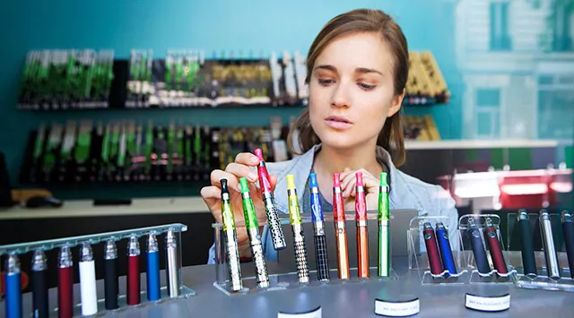 A vape store owner examining a red vape in a well-organized store. Various products are on the shelves in the background.