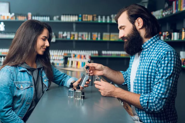 Man showing a vape bottle to a woman in a vape store with shelves of products in the background.