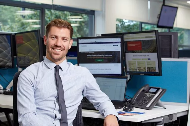 Smiling insurance broker at a desk with computer screens displaying websites and a phone on his desk.