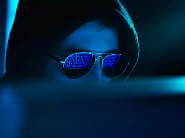 Hacker in a dark hoodie working on a laptop in a dimly lit room with code reflected on his black sunglasses.