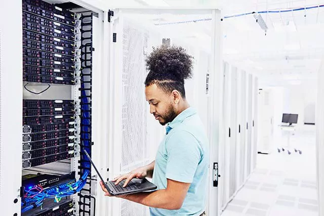 A person with curly hair in a bun is standing in a server room, using a laptop. They are in front of a server rack filled with various networking equipment and cables. The room is well-lit, with multiple server cabinets visible.