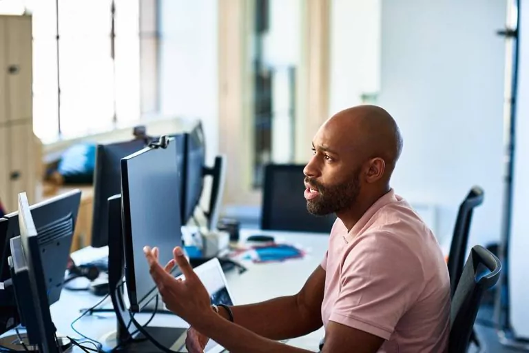Man gesturing while talking at a desk facing a turned-off monitor.