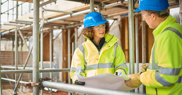 Two construction workers in yellow jackets and blue helmets discuss plans at a site covered by scaffolding.